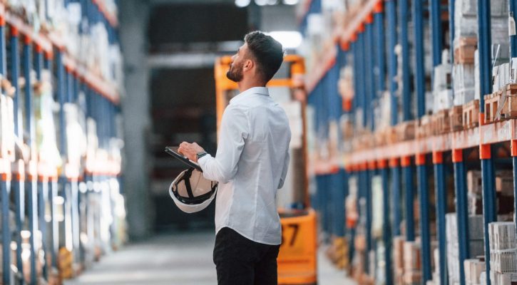 With tablet in hands. Storage worker is in the warehouse with bunch of products.
